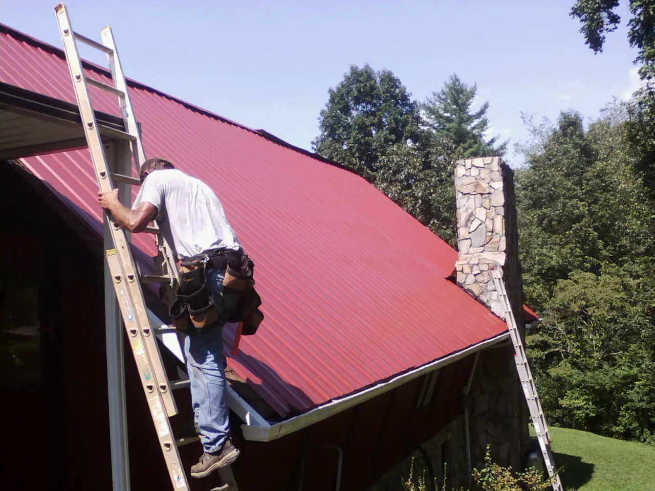 Roofer on a ladder beside a red metal roof and stone chimney — Soffit & Fascia Repair by Roofing Brothers in Broken Arrow